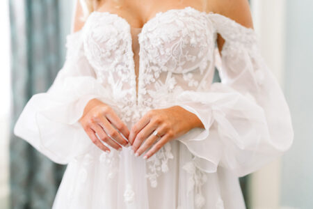 Bride in off-shoulder white lace wedding dress with sheer sleeves, hands together showing a simple ring.