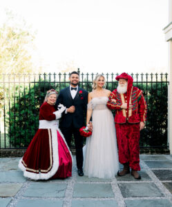Four adults smile outdoors in formal attire—burgundy gown, tuxedo, wedding dress with bouquet, and festive red-gold outfit—by an iron fence.