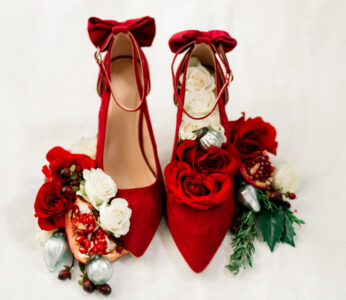 Red high heels with bows, surrounded by roses, pomegranate halves, pine greenery, berries, and silver ornaments on white.