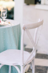 White wooden chair by a round table with light blue textured tablecloth, glassware, and cutlery.