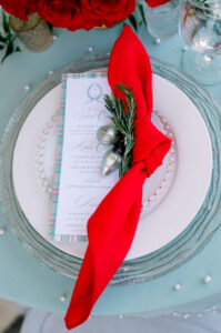 Elegant table setting with menu, red napkin and rosemary on white plate, pearl-accented blue cloth, and red flowers behind.