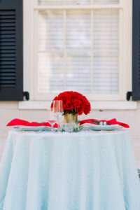 Round table with blue pearl-dotted cloth, red napkins, glassware, red rose centerpiece by a window with black shutters.