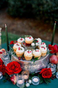 Cupcakes with white frosting and red berries on a silver stand, surrounded by roses, pomegranates, candles, and greenery.