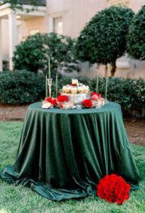 Outdoor round table with dark green cloth, candles, red roses, desserts; bouquet on grass, bushes and building behind.