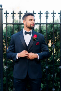 Man in black tuxedo with red rose boutonniere adjusts jacket by wrought iron fence and greenery, looking to the side.