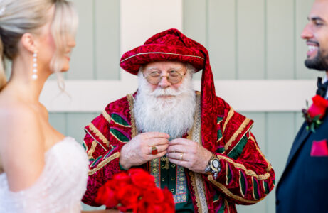 A man in a red, gold, and green festive costume with a long white beard stands between a smiling bride and groom.