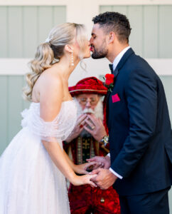 Bride and groom holding hands lean in for a kiss as Santa Claus behind them covers his mouth in surprise.