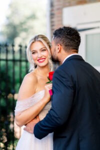 Bride in an off-shoulder dress with red rose, groom in dark suit whispering near black fence and brick wall outdoors.
