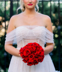Bride in white off-shoulder dress holds red roses, standing by a wrought iron fence with greenery behind her.
