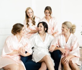 Five women in satin robes, one white and four pink, laugh together on a dark blue couch before their New Orleans wedding.
