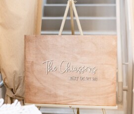 Wooden sign on easel with “The Chiassons EST. 05.27.23” in white script, in front of stairs with striped curtains.