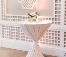 Round cocktail table with white cloth, florals, and candles on wood floor by a white lattice wall—ideal for Louisiana weddings.