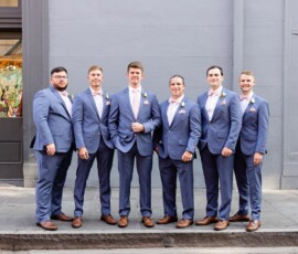 Six men in blue suits and brown shoes pose on a sidewalk against a gray wall at a New Orleans wedding.