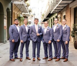 Six men in light blue suits and brown shoes pose together on a historic city street, offering stylish wedding inspiration.