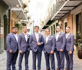 Six men in light blue suits and brown shoes pose on a sunny Louisiana street; the groom stands slightly ahead, smiling.