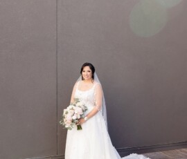 Bride in white dress with pink and white bouquet stands on wooden floor by gray wall, perfect Louisiana wedding inspiration.