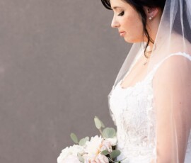 Bride in white lace dress and veil holds pastel bouquet, looking down thoughtfully against a plain gray background.