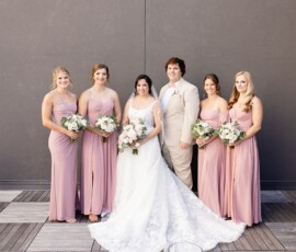 Bride, groom, and four bridesmaids in pink pose with bouquets before their New Orleans wedding against a gray wall.