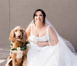 Bride in white dress smiles next to brown dog with floral collar, both posed by a gray wall—charming New Orleans wedding moment.