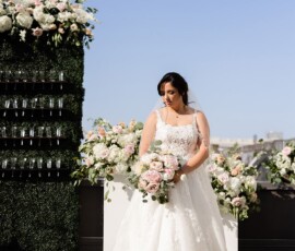 Bride in white gown holding bouquet outdoors amid flowers and greenery under blue sky at a Louisiana wedding.