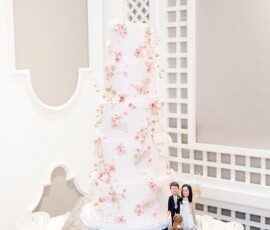 White wedding cake with pink flowers on a silver stand, bride and groom figurines, dog, and candles on a round table.