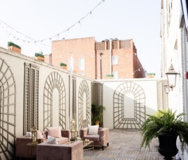 Charming New Orleans patio with wooden floors, string lights, trellis walls, plush chairs, potted ferns, and brick building views.