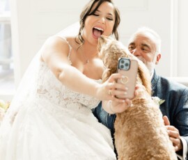 Bride in white dress takes a joyful selfie with a man and fluffy dog licking her face at a bright Louisiana wedding.