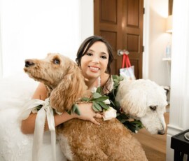 Bride in white dress hugs two large dogs with flower collars, smiling in a bright room before her Louisiana wedding.