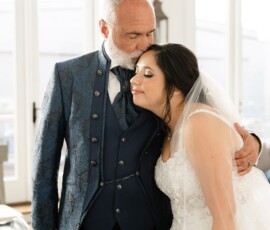 An older man in a blue suit hugs and kisses a smiling bride in white lace at a sunlit Louisiana wedding.