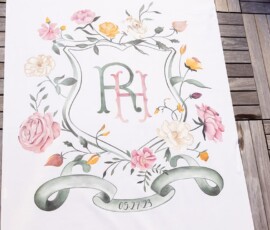 Elegant floral crest with RH initials, pink, yellow, peach flowers, and “05.27.23” on green ribbon at New Orleans wedding.