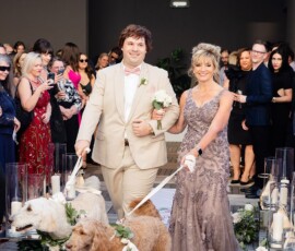 Couple in formal attire walk two flower-collared dogs down candlelit aisle at romantic New Orleans wedding.