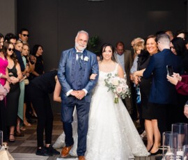 Bride in white gown and bouquet walks with older man in blue suit down aisle at elegant Gulf South wedding, guests smiling.