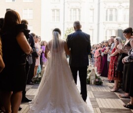 Bride in a white gown and veil walks an outdoor aisle with an older man, as guests watch at a New Orleans wedding.