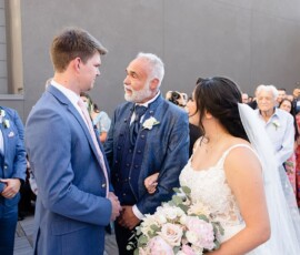 Bride with bouquet stands by older man, handing her to groom at outdoor Louisiana wedding as guests watch.