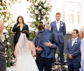 Bride in white gown with groom hugging older man at floral altar, groomsmen in blue suits, and officiant at Louisiana wedding.