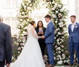 Bride and groom hold hands by a floral arch as officiant reads at outdoor Louisiana wedding; formal attire, flowers, greenery.