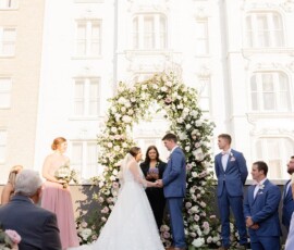 Bride and groom hold hands under a floral arch at their outdoor Louisiana wedding, with formal party and ornate building behind.