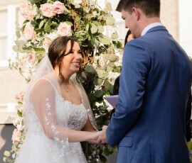 Bride and groom smiling and holding hands by a floral arch at their outdoor Louisiana wedding ceremony.