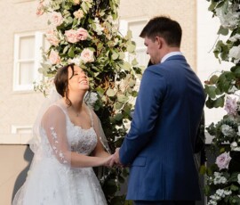 Bride and groom hold hands, smiling at their outdoor Louisiana wedding amid pink and white rose floral arches.