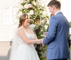 Bride in white dress and groom in blue suit smile under a floral arch at their outdoor New Orleans wedding ceremony.