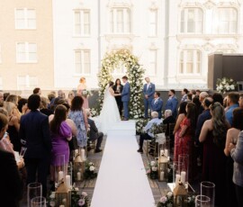 Bride and groom under a floral arch at outdoor Louisiana wedding, guests seated along a white aisle.