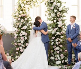 Bride and groom kiss under a flower arch at their Gulf South outdoor wedding, surrounded by guests and attendants.