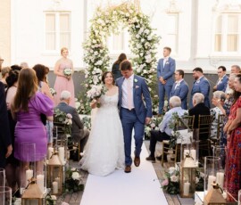 Bride and groom smile walking down the aisle at an outdoor Gulf South wedding with floral decor and a white arch.