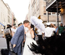 Bride and groom kiss in a New Orleans wedding parade, surrounded by musicians and fans with black and white feathers.