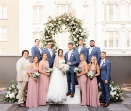 Bride and groom with wedding party under floral arch at a scenic Louisiana outdoor wedding, all smiling.