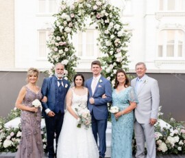 Bride and groom under a floral arch at their outdoor New Orleans wedding, smiling with four adults, white architecture behind.