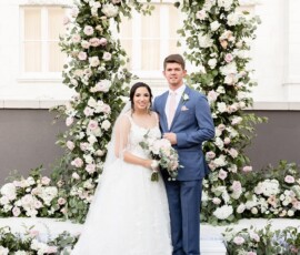 Bride in white gown and groom in blue suit smile before a pink and white floral arch at a romantic Louisiana wedding.