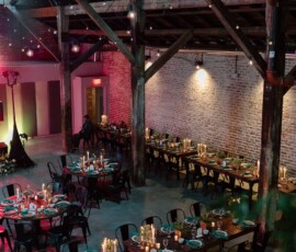 Rustic industrial event space featuring exposed brick, wooden beams, long tables with candles and greenery, and cozy lighting.