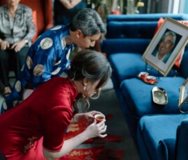 Couple in traditional attire kneel and present tea during a cozy Louisiana wedding ceremony, observed by family in a living room.