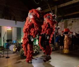 Red lion dancers perform at an indoor Gulf South wedding as guests watch, with instruments and a neon sign in festive lighting.